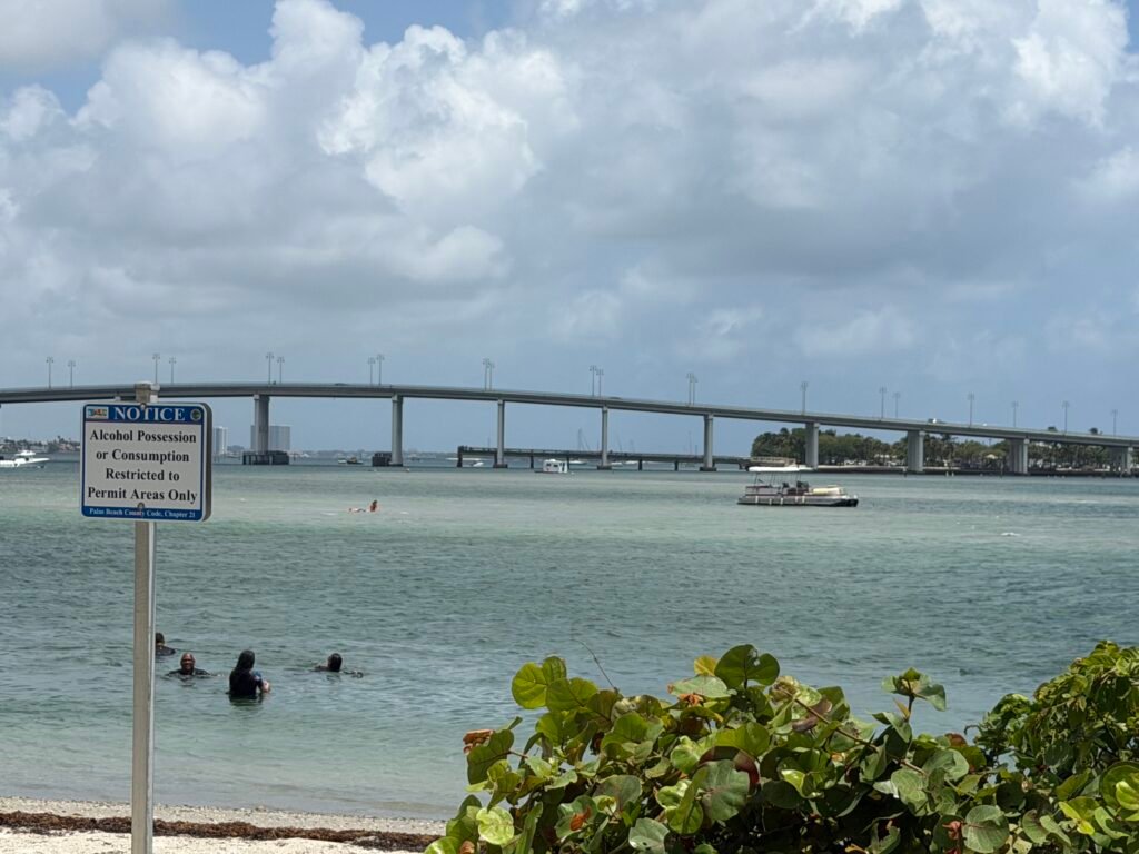 Alcohol restriction sign on a Peanut Island beach, with swimmers and the Blue Heron Bridge behind it
